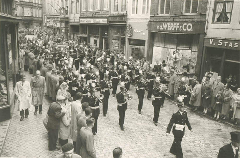 Het muziekkorps van de mijnen loopt door een straat in Maastricht. Aan de kant staan toeschouwers te kijken en te luisteren