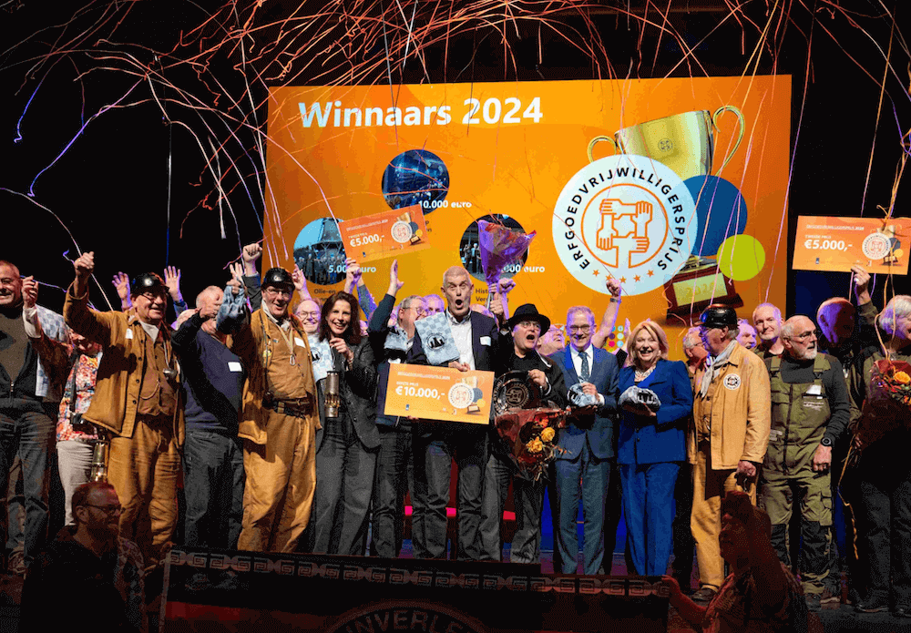 Podium met daarop het winnende team van de Erfgoedvrijwilligersprijs, te midden van allerlei andere erfgoedvrijwilligers. Een van de winnende teamleden draagt een bord met daarop de prijs: 10.000 euro