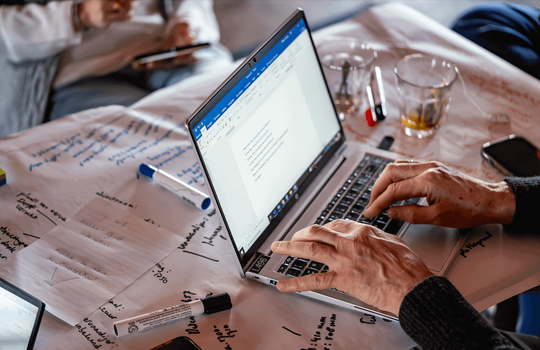 Handen boven een laptop op een tafel vol beschreven vellen papier