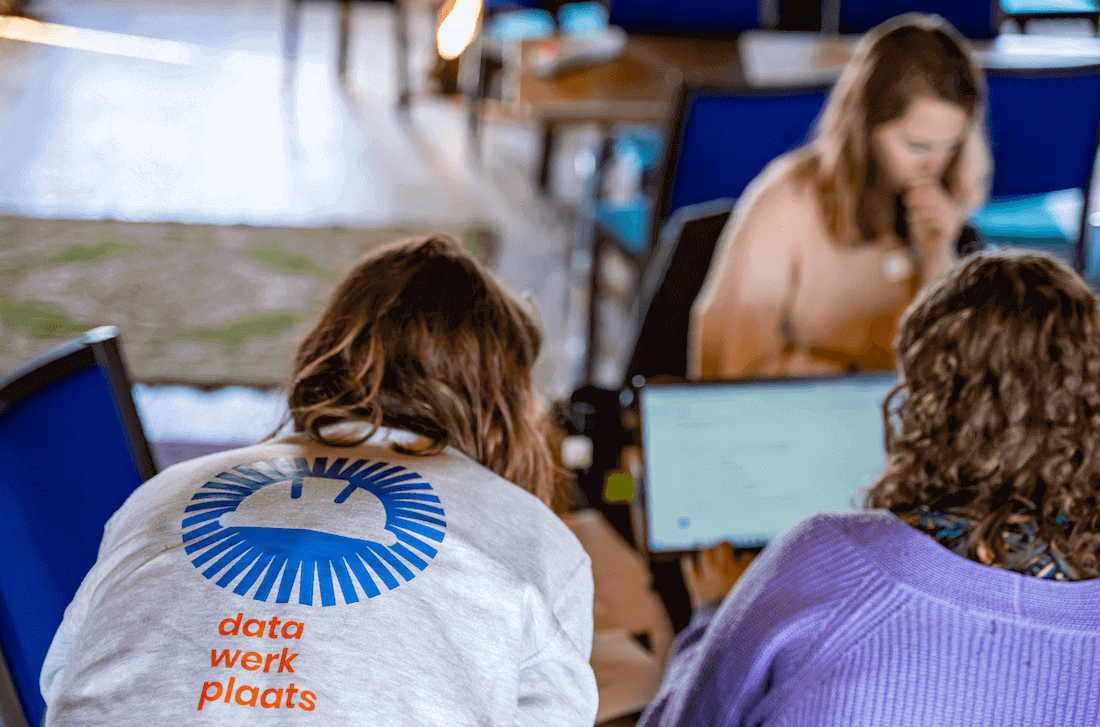 Twee deelnemers achter laptops krijgen hulp van een begeleider met 'datawerkplaats' op de rug
