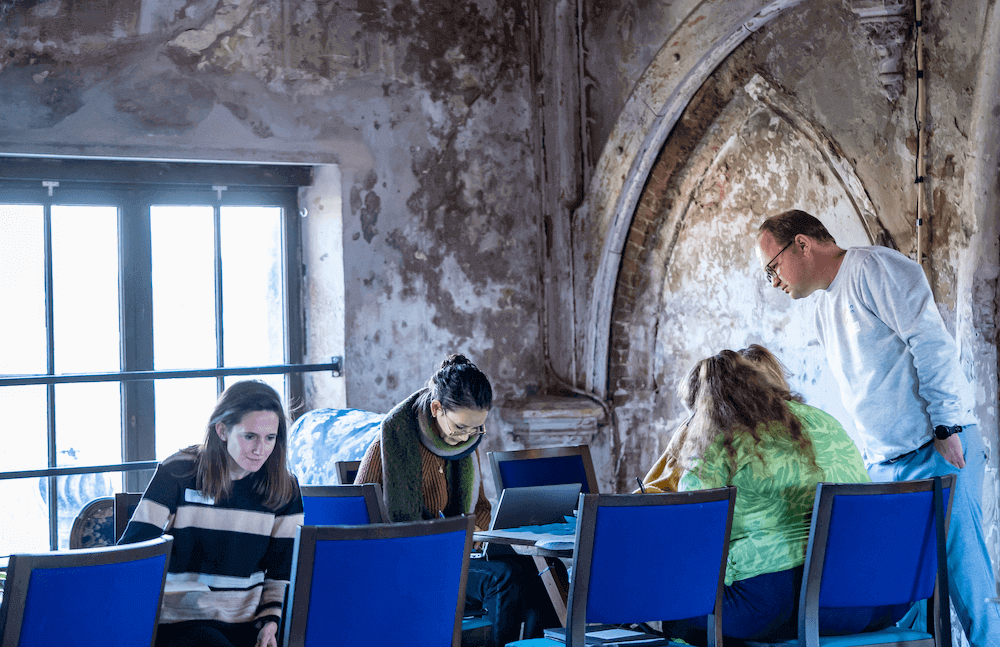 Foto van een groep mensen die aan een tafel achyter laptops zitten te werken. De omgeving is monumentaal met afgebladderde muren. Een workshopbegeleider kijkt toe