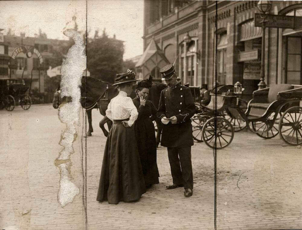 Voor het station in Arnhem vragen twee dames in lange japonnen informatie aan een spoorwegmedewerker