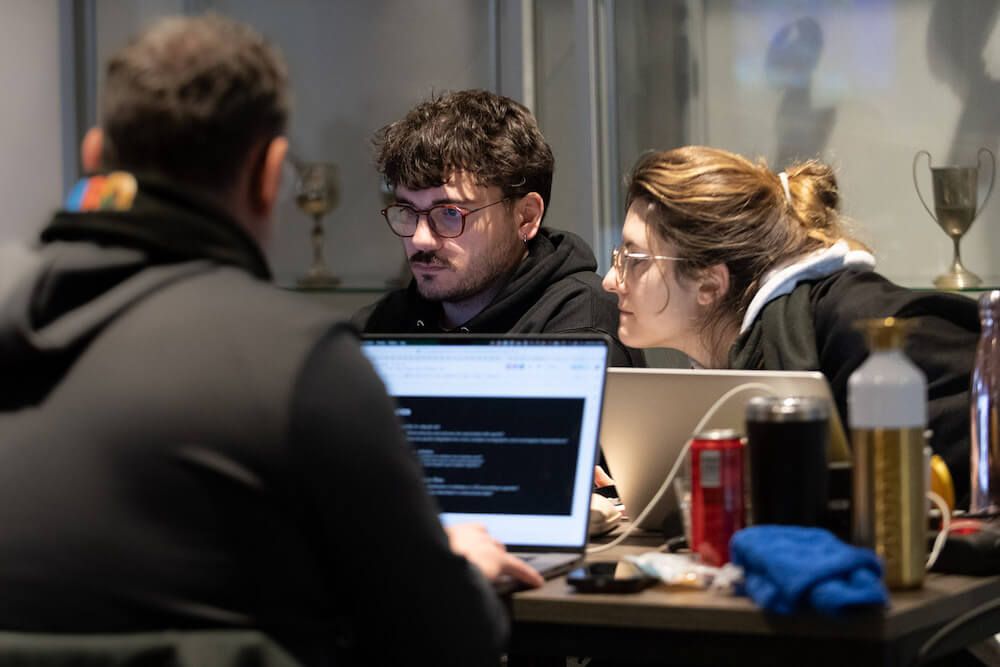 Foto van drie mensen die achter laptops zitten te werken aan een tafel met allerlei spullen zoals waterfles, coca-colablikje en andere spullen