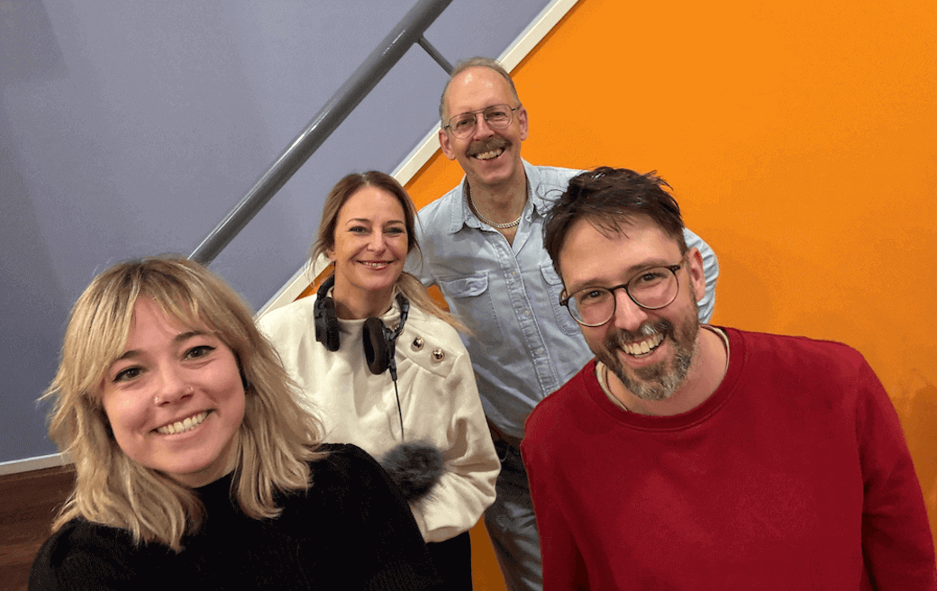 Foto van 4 staande mensen die meewerken aan de podcast. Ze staan achter een trap met felle oranje wand